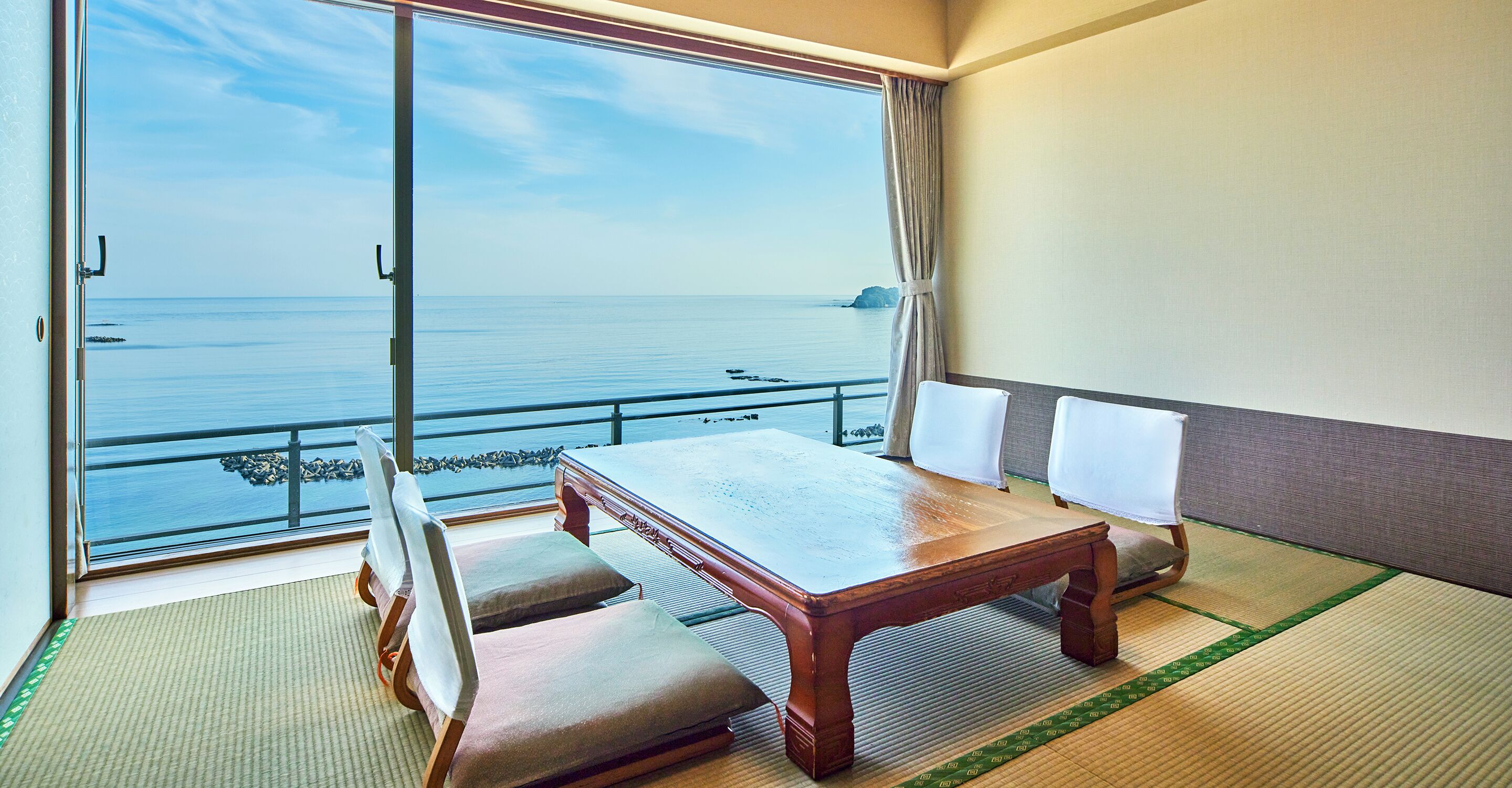 Een kamer met een houten tafel en stoelen, een groot raam met uitzicht op zee, en tatami matten op de vloer.