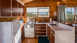 A compact kitchen with wooden cabinets, a stove, a sink, and a window with blinds. - Beach House on the Ridge - Kaiteriteri House (Little Kaiteriteri)