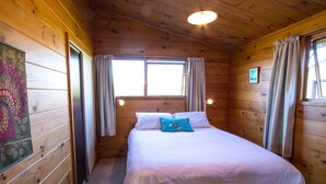A wooden cabin bedroom with a bed, curtains, and a window. - Beach House on the Ridge - Kaiteriteri House (Little Kaiteriteri)