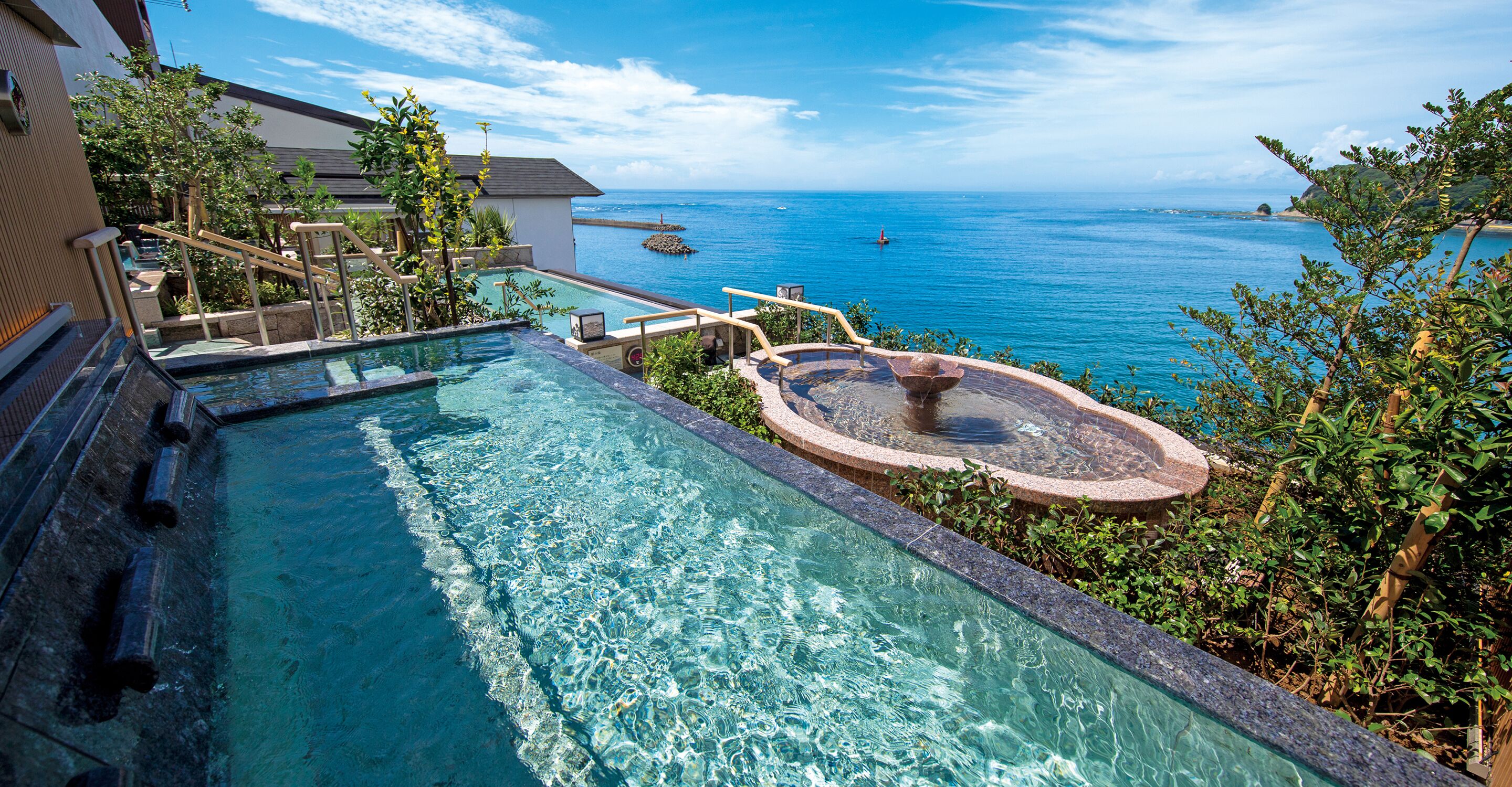 A pool with a view of the sea, a hot tub, and a wooden deck with railings.