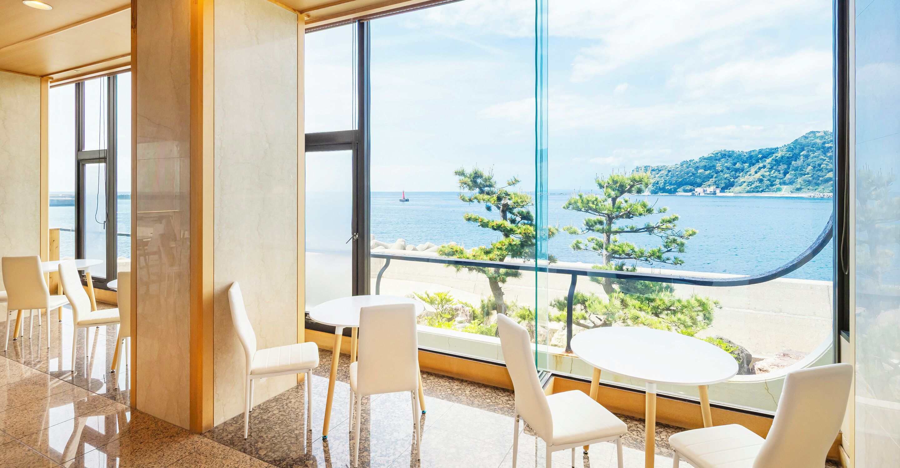A modern interior with large windows offering a view of the sea and a tree-lined path.
