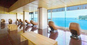 A room with reclining chairs and a wooden bench, large windows with a view of the sea.