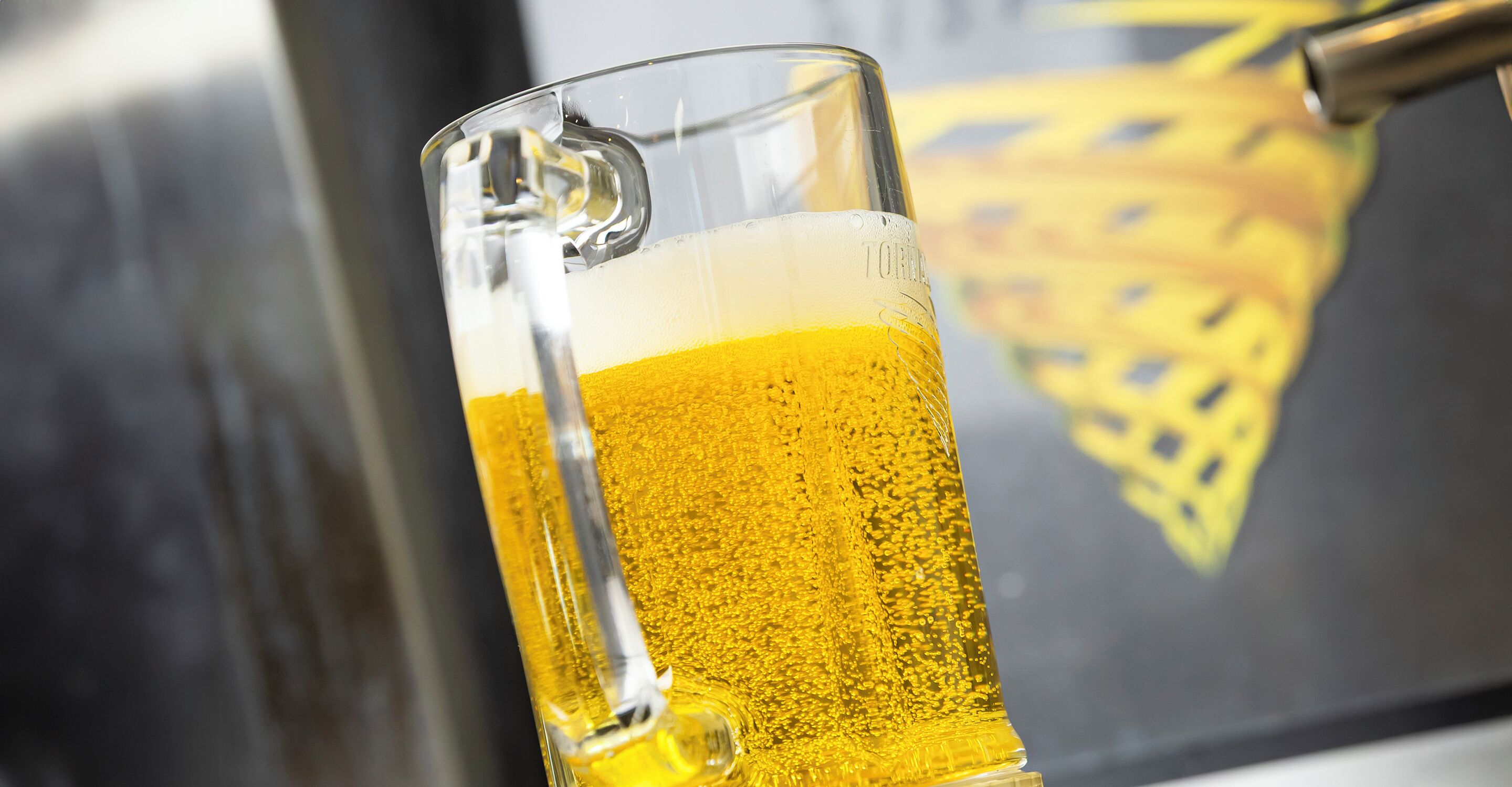 A clear glass mug filled with a golden liquid, likely beer, with a tap attached to the side.