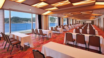A conference room with tables and chairs arranged for a meeting, large windows offering a view of the sea, and a ceiling with wooden beams.