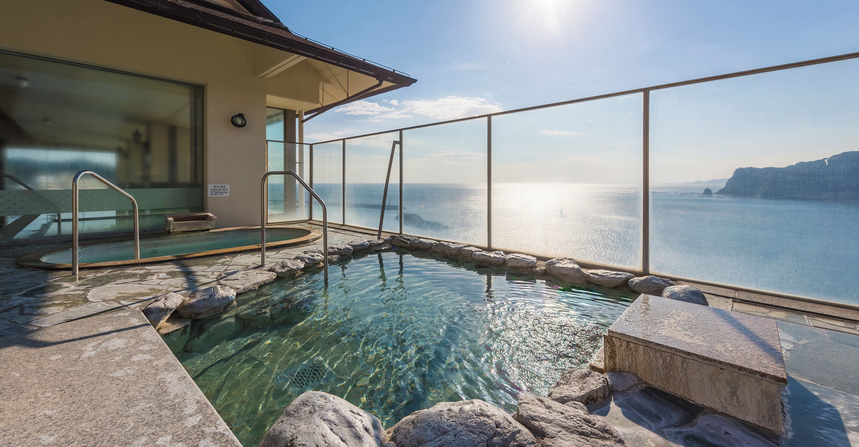 An infinity pool with clear water, surrounded by rocks and a glass railing, overlooking a body of water and distant land.
