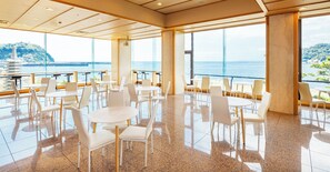A spacious indoor dining area with multiple tables and chairs, large windows offering a view of the sea and a distant island. - Mikazuki Sea-Park Hotel Awa Kamogawa (Kamogawa)