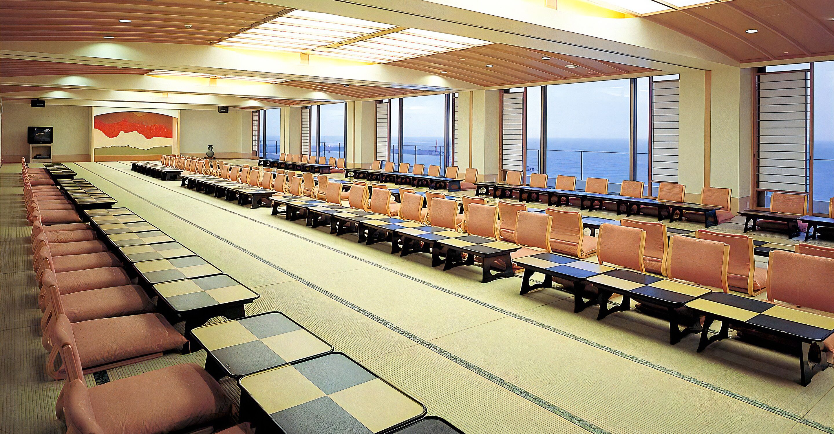 A conference room with rows of tables and chairs, large windows, and a view of the sea.