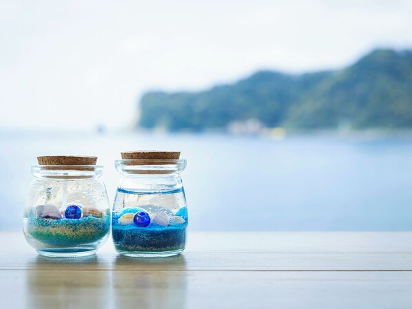 Two glass jars with cork lids containing blue sand and seashells, placed on a wooden surface with a blurred coastal background. - Mikazuki Sea-Park Hotel Awa Kamogawa (Kamogawa)