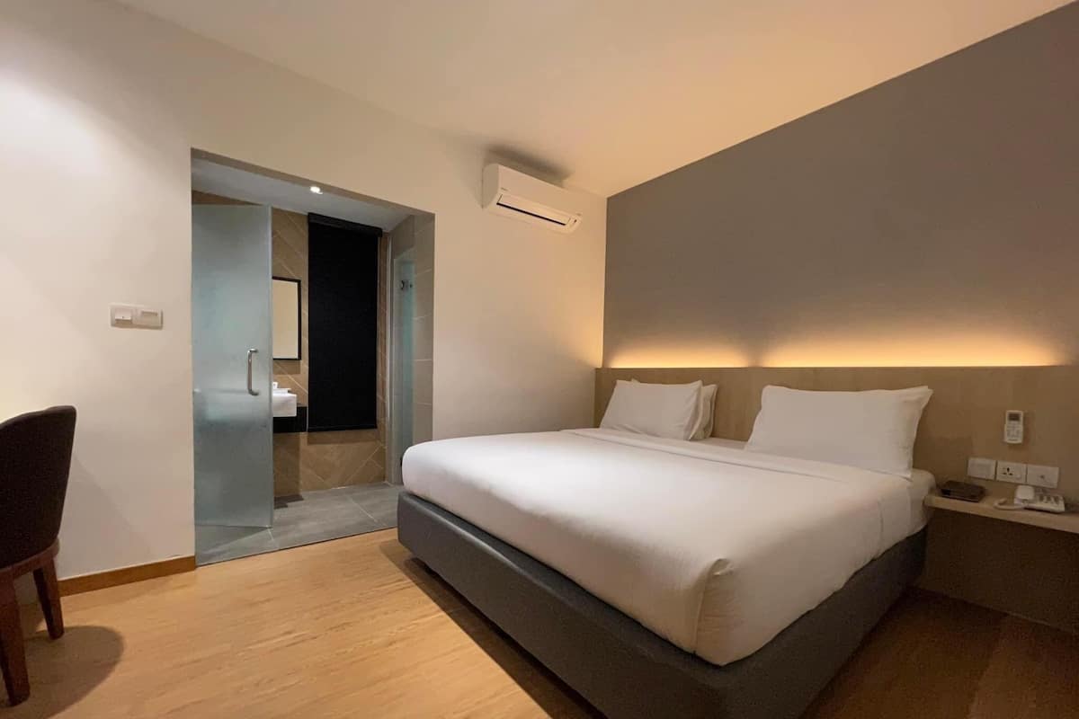 A modern hotel room with a large bed, a bedside table with a lamp, a chair, and a bathroom visible through an open door.