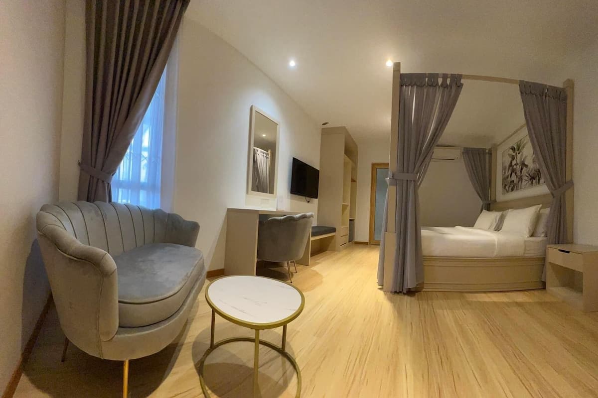 A modern hotel room with a grey sofa, a small round table, a bed with curtains, a TV, and a painting on the wall.