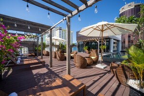 Una terraza de madera con mobiliario de mimbre, sombrillas y una zona de piscina.