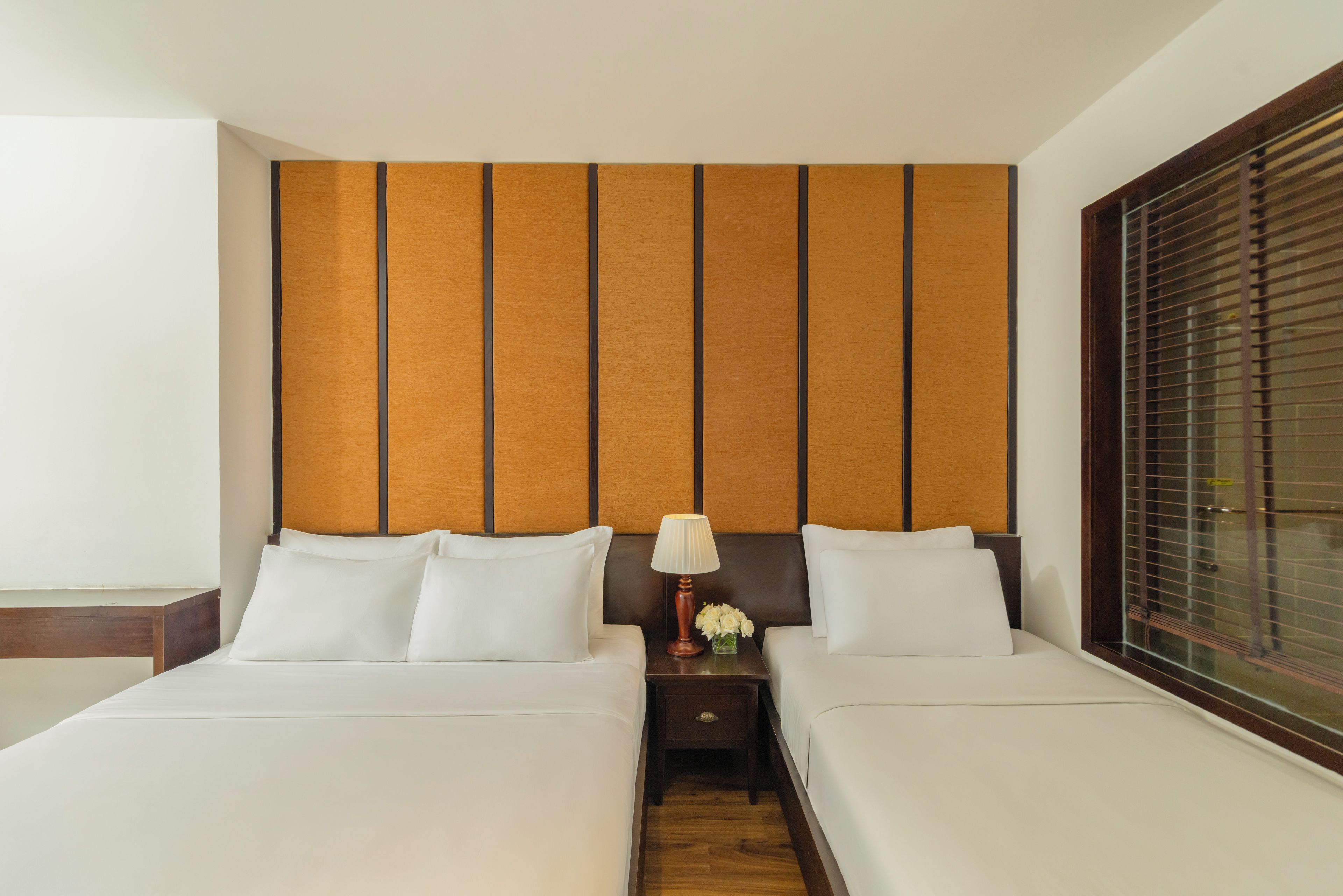 A hotel room with two beds, a nightstand with a lamp, and a window with blinds.