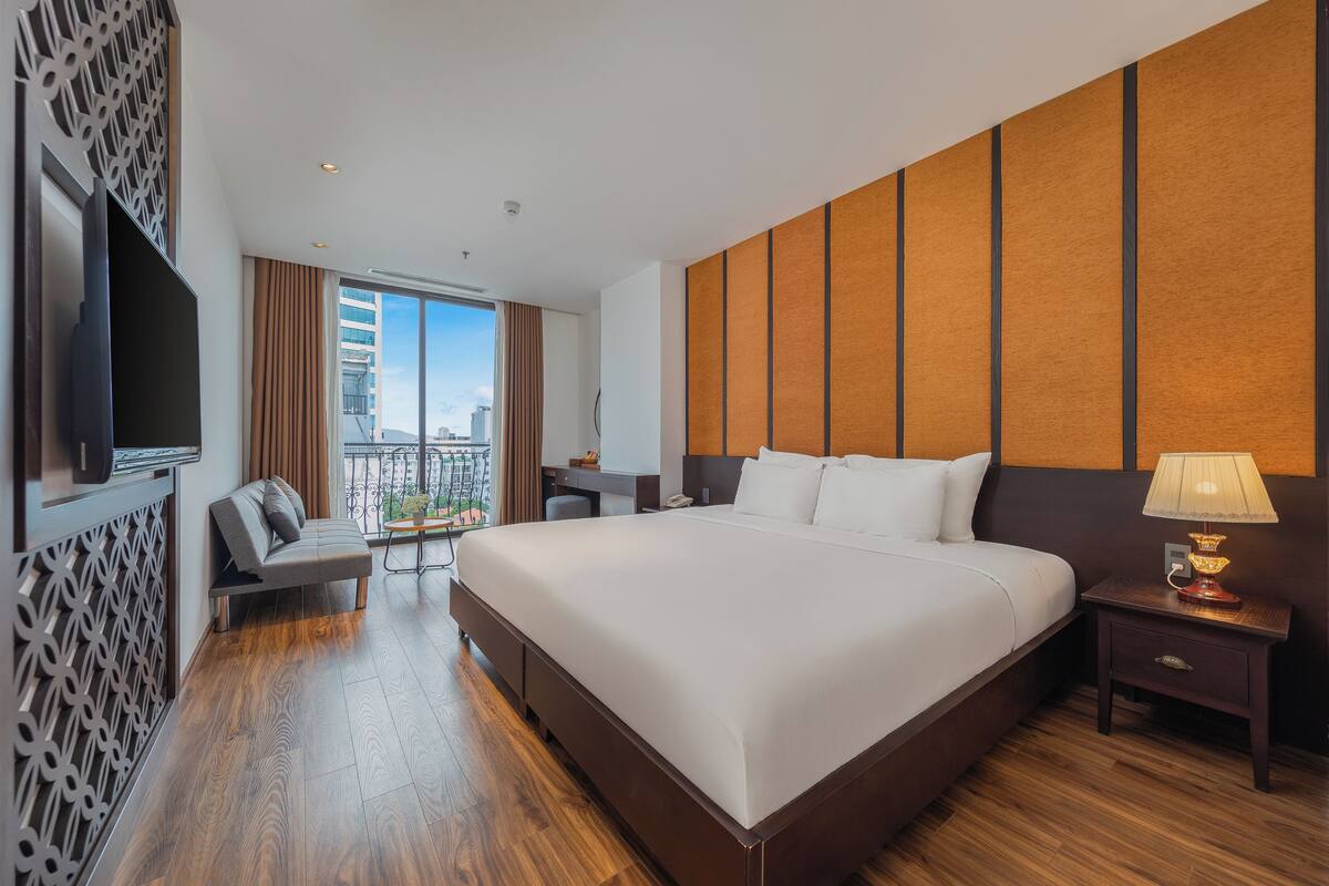 A modern hotel room with a large bed, a desk, a TV, and a balcony with a city view.