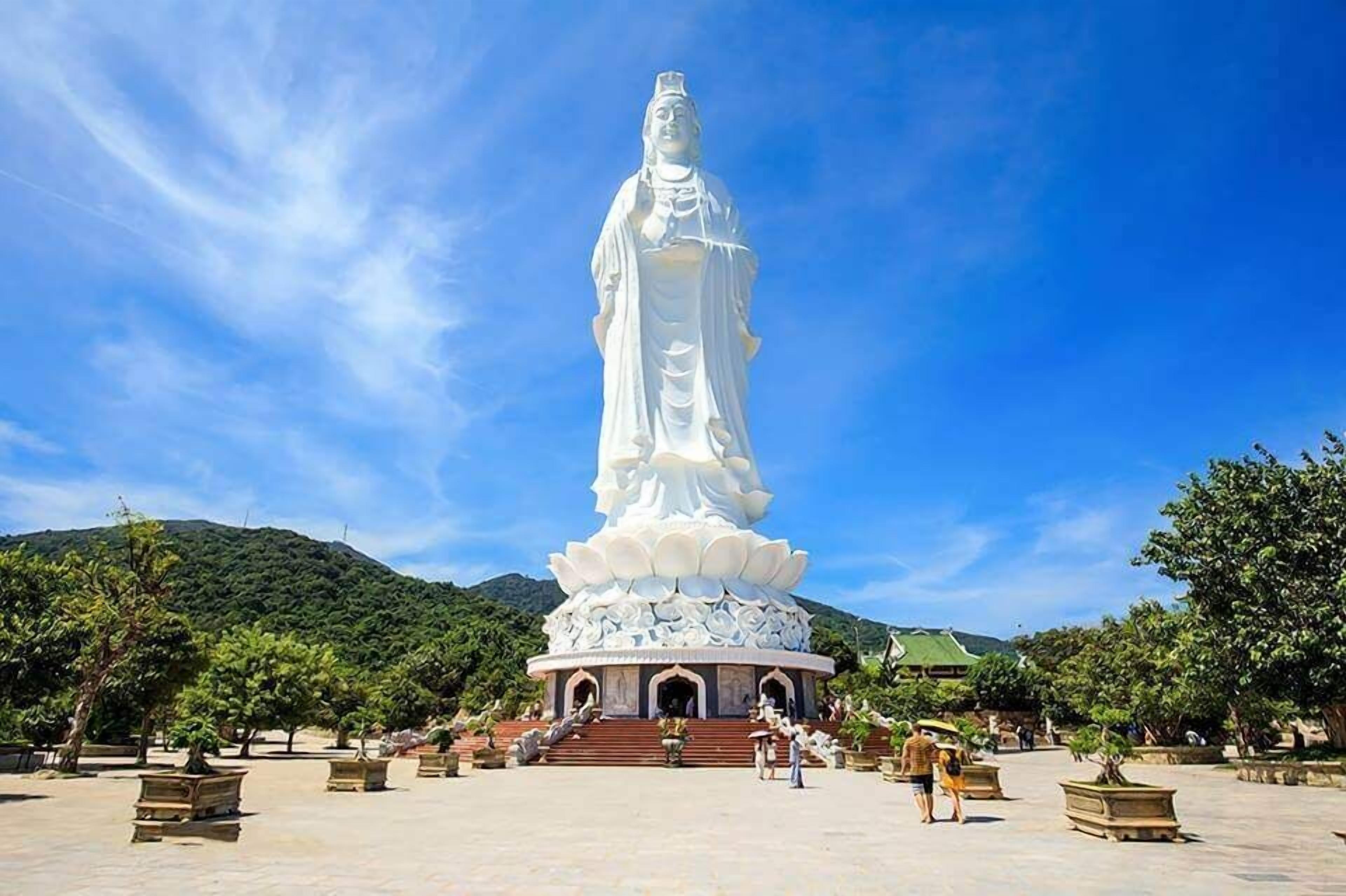 A large white statue with a lotus base, surrounded by a paved area with benches and trees.