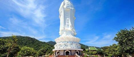 A large white statue with a lotus base, surrounded by a paved area with benches and trees.