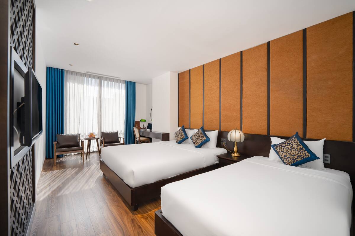 A hotel room with two beds, a TV, a desk with a lamp, and a view of the outdoors through large windows.