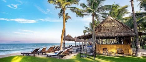 Un resort frente a la playa con sillones de descanso, palmeras y una cabaña de techo de paja.