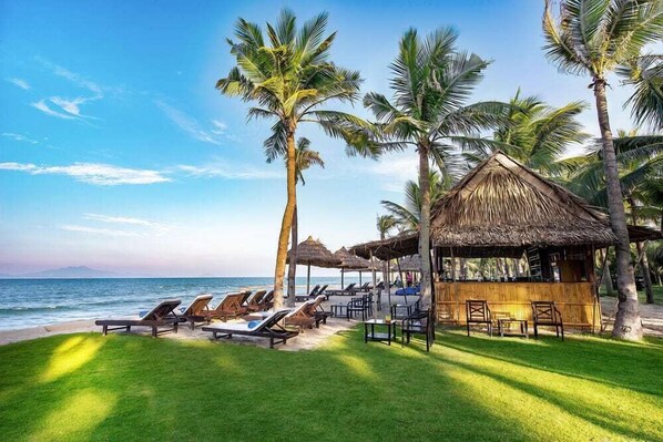 Un resort frente a la playa con sillones de descanso, palmeras y una cabaña de techo de paja.