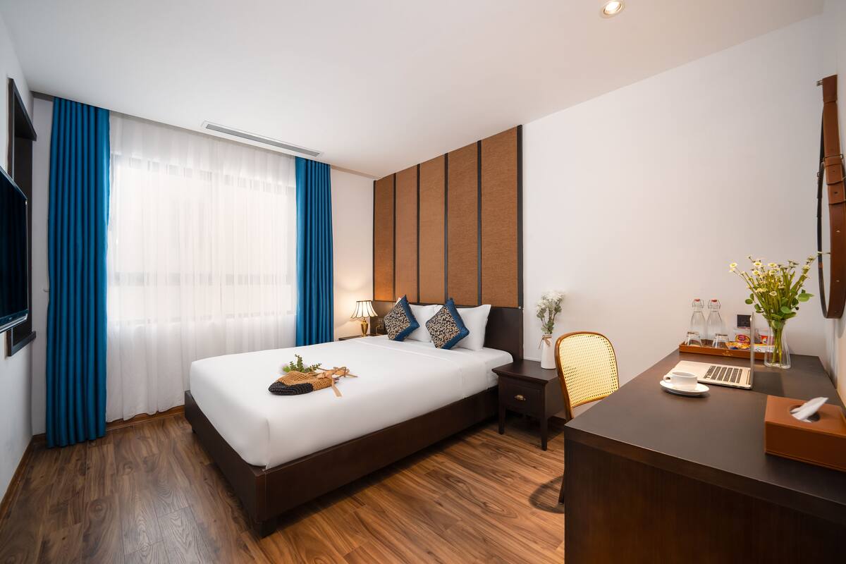 A modern hotel room with a large bed, wooden flooring, a desk with a laptop, a chair, and a bedside table with a lamp.