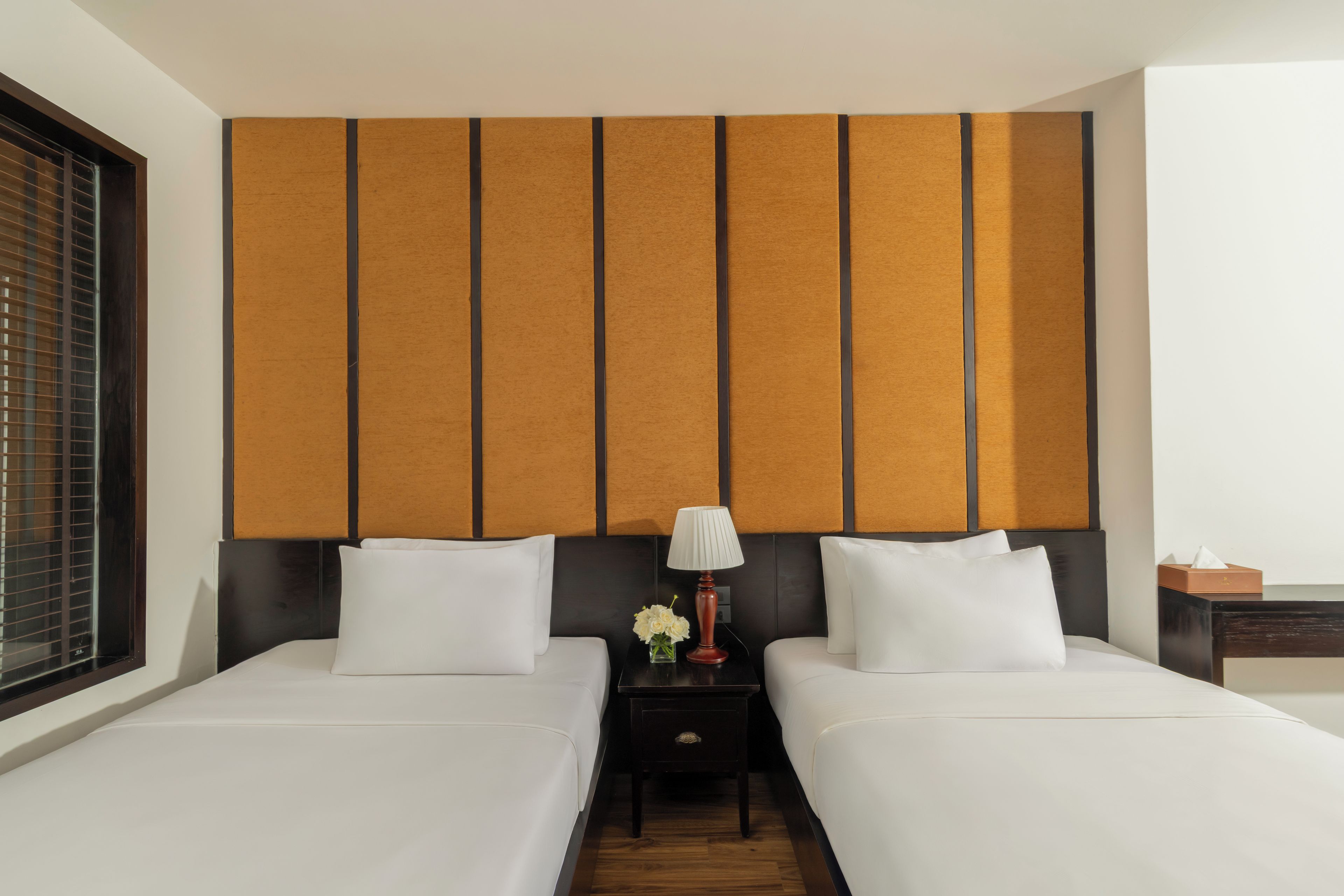Habitación de hotel con dos camas, una mesita de noche con lámpara y una ventana con persianas.