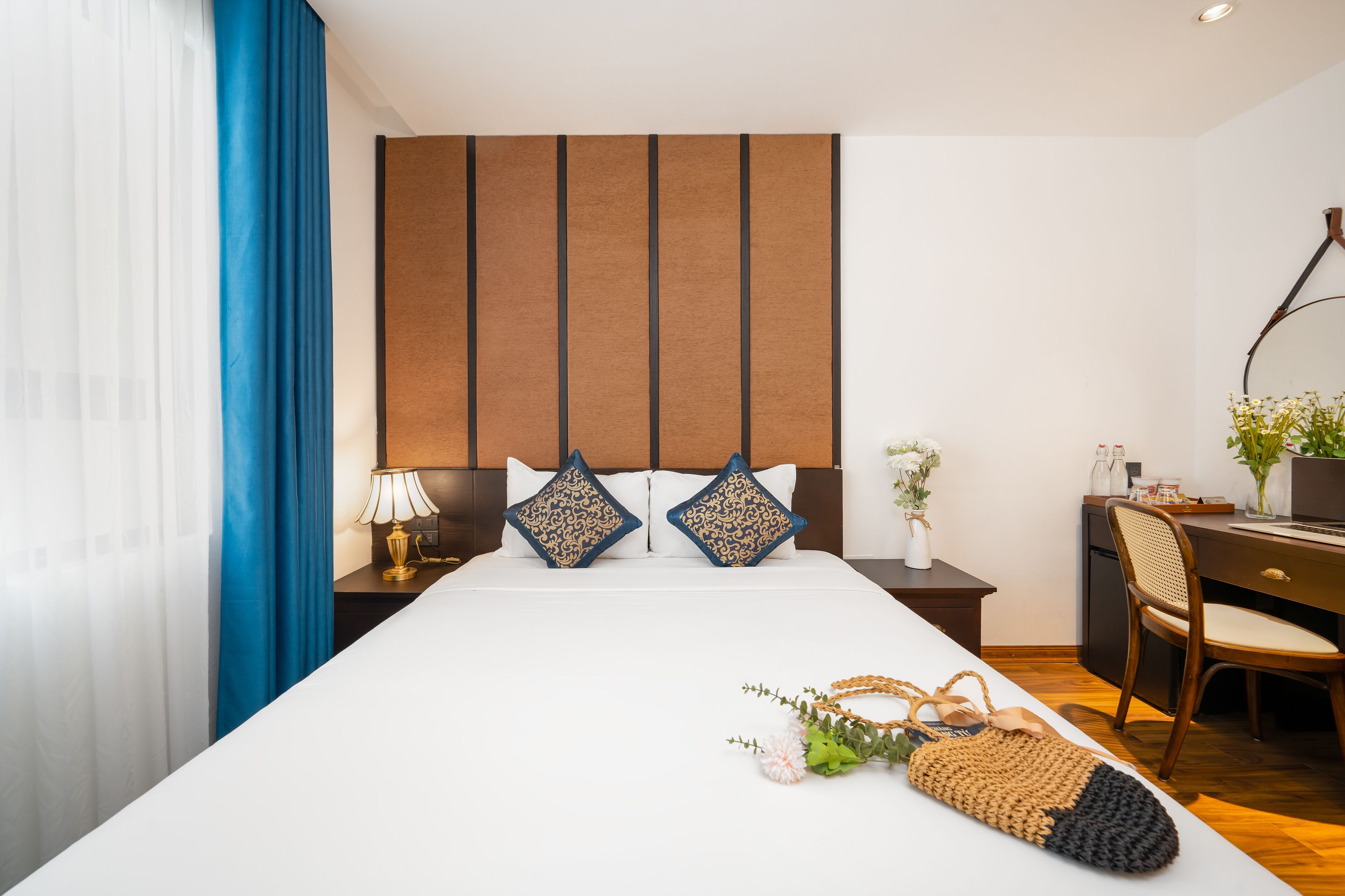A hotel room with a large bed, two decorative pillows, a desk with a chair, a lamp, and a vase with flowers.