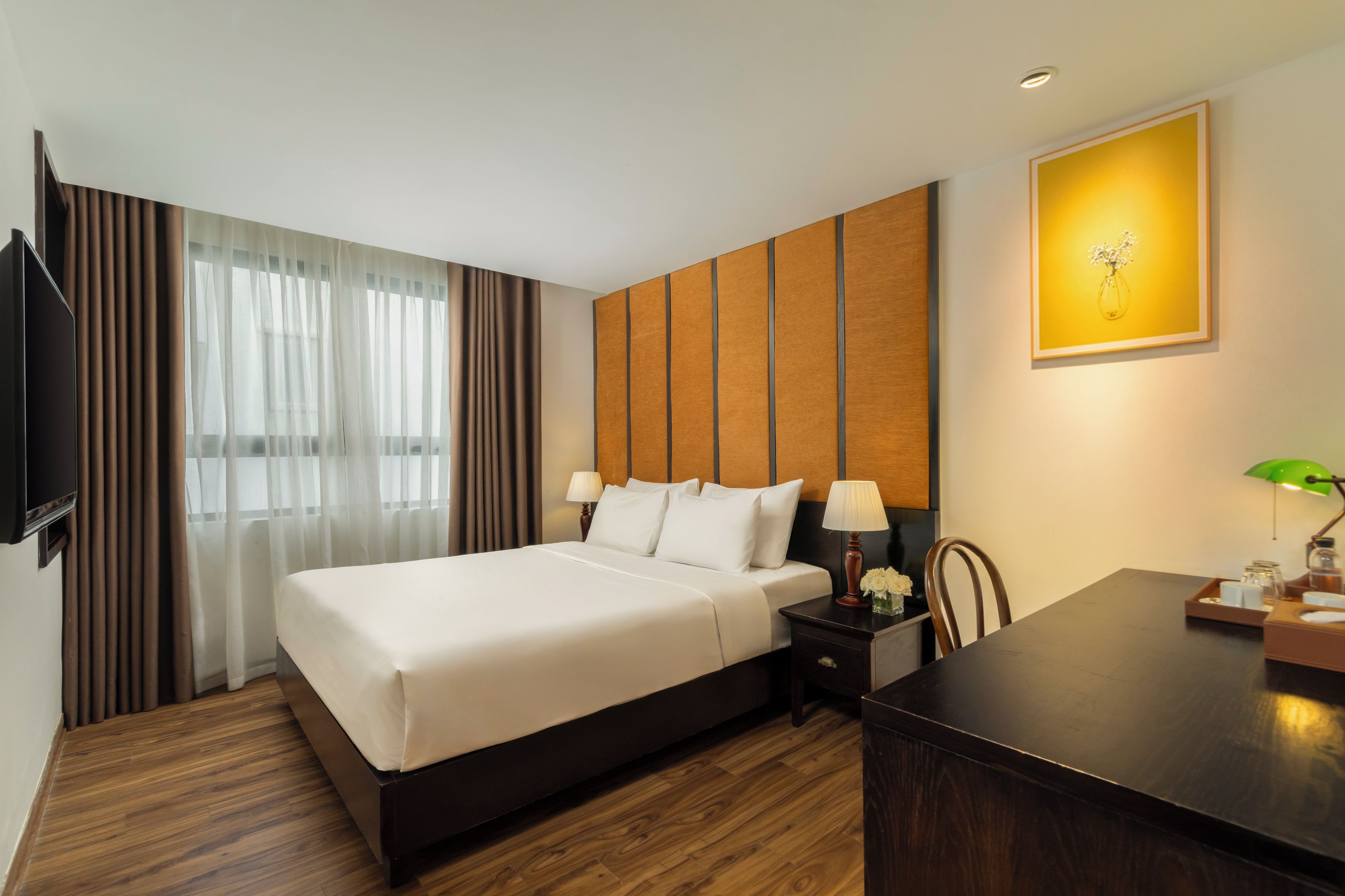 A hotel room with a large bed, a desk with a lamp, a television, and a painting on the wall.