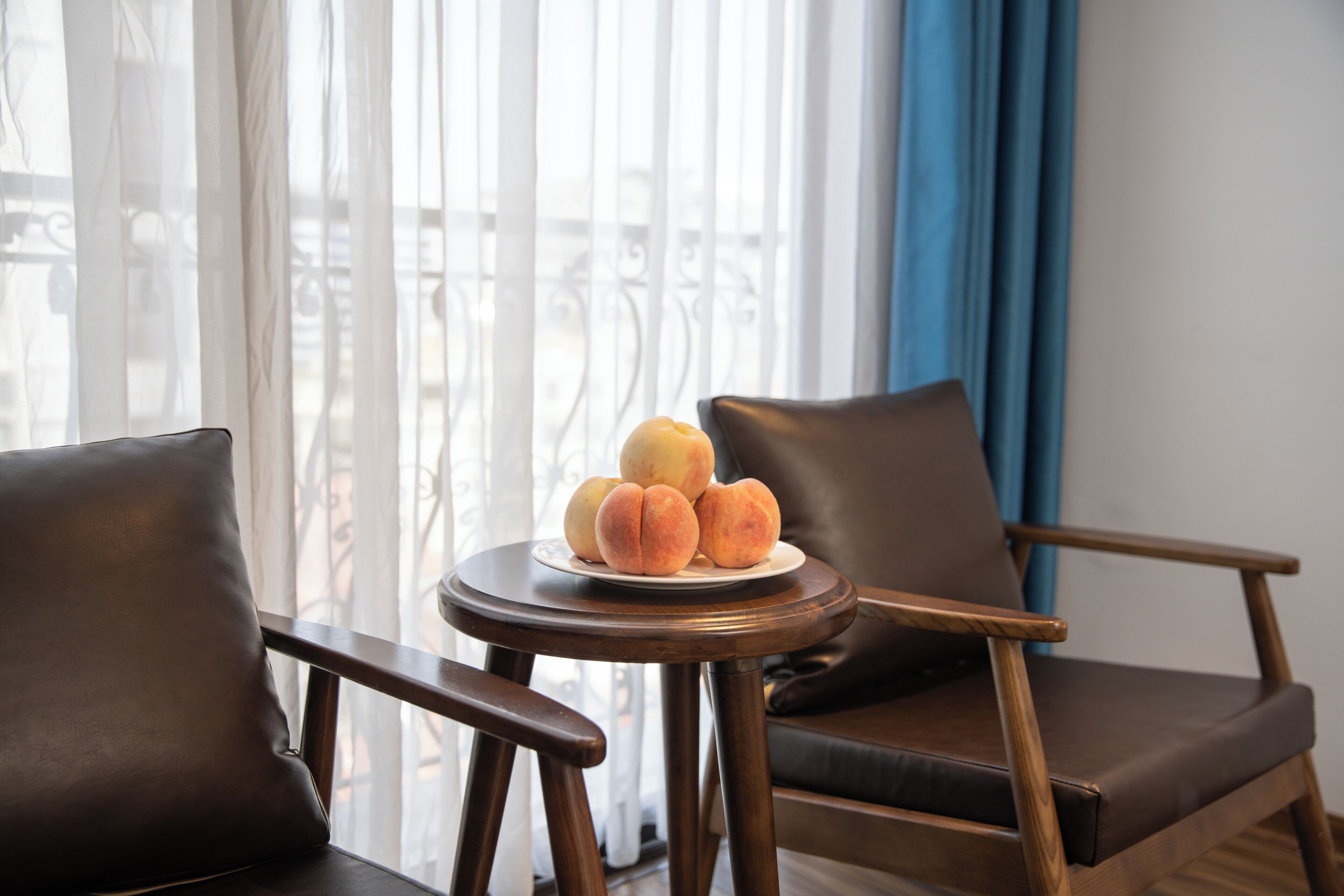 Una habitación con vistas a un balcón, una mesa redonda de madera con un plato de melocotones y dos sillones de cuero.