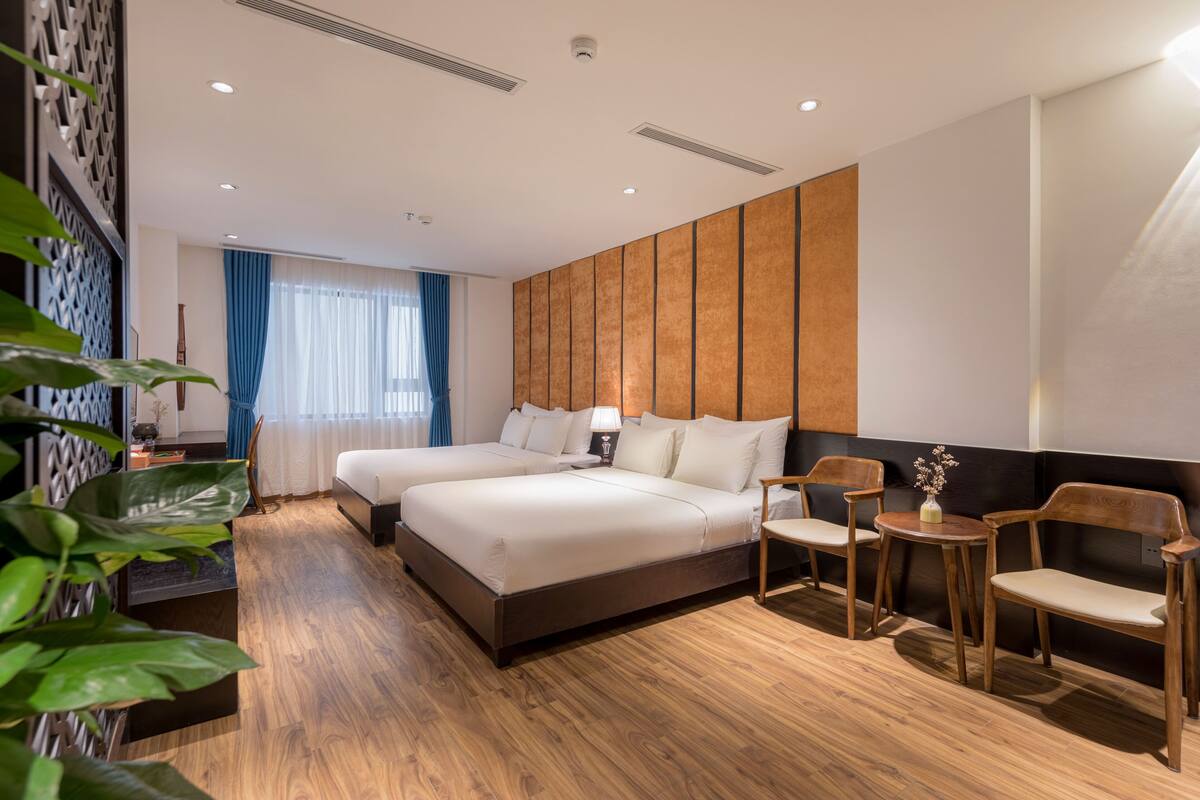A hotel room with two beds, wooden flooring, a small table with chairs, and a decorative wall panel.