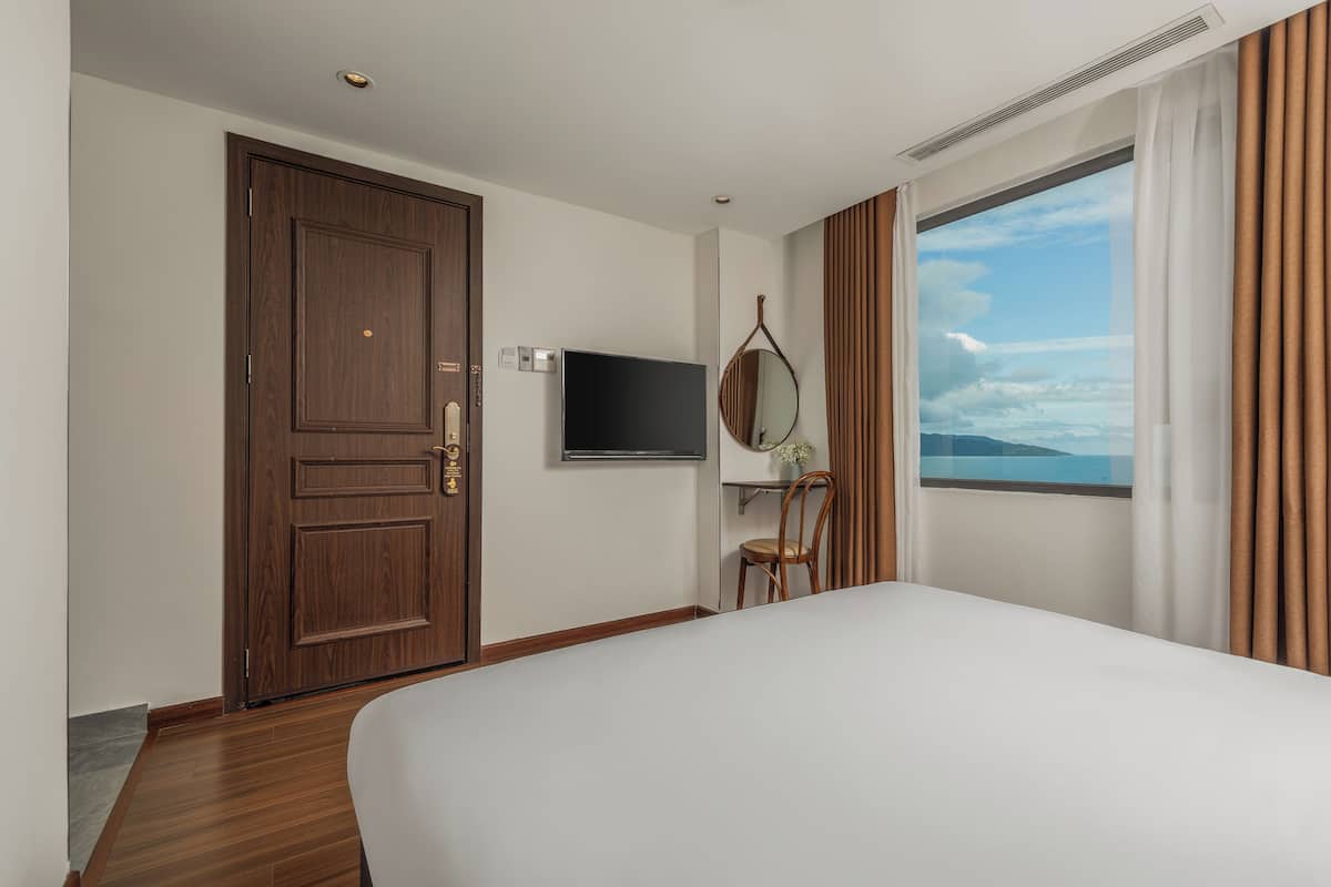 A hotel room with a large bed, a wooden door, a flat-screen TV, a chair, a small table, a window with curtains, and a view of the sky.