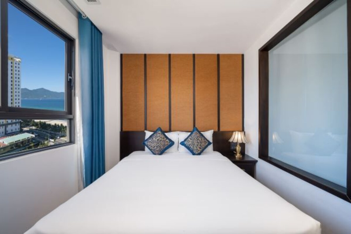 A hotel room with a large bed, a headboard with vertical panels, a window with a view of buildings and mountains, and a nightstand with a lamp.