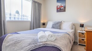 Cama arrumada com uma toalha enrolada, abajur e mesa de cabeceira de madeira com cestos.