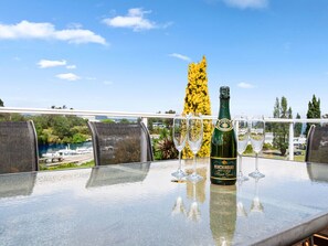 A table with a bottle of Hinchinbury wine and three glasses on a balcony overlooking a scenic view. - Central Riverside Retreat - Taupo Holiday Home (Taupo Central)