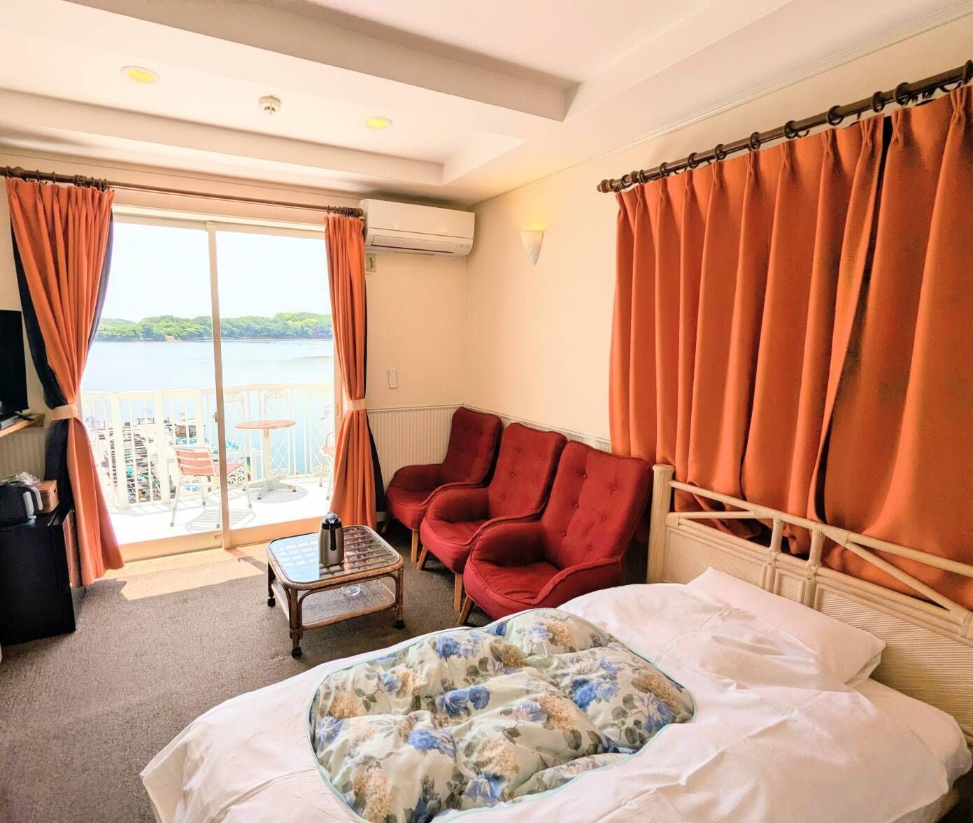 A hotel room with a bed, two red armchairs, a small table, and a view of the sea through a large window.