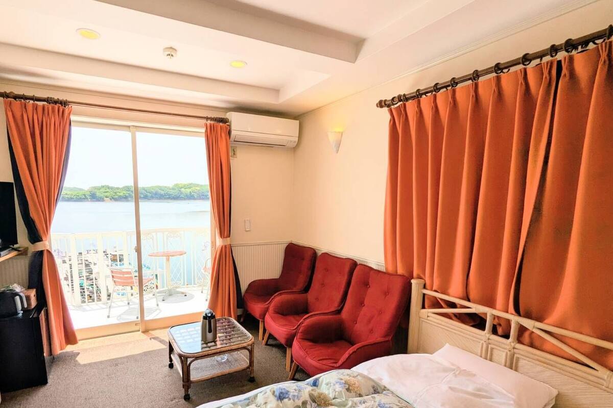 Habitació d'hotel amb un llit, dues butaques vermelles, una taula petita i vista al mar a través d'una finestra gran.
