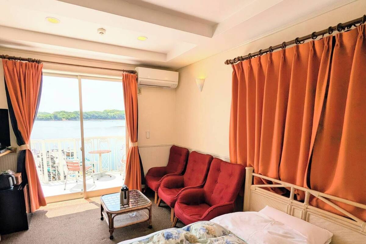Habitació d'hotel amb un llit, una butaca vermella, una taula petita i vista al mar a través d'una finestra gran.