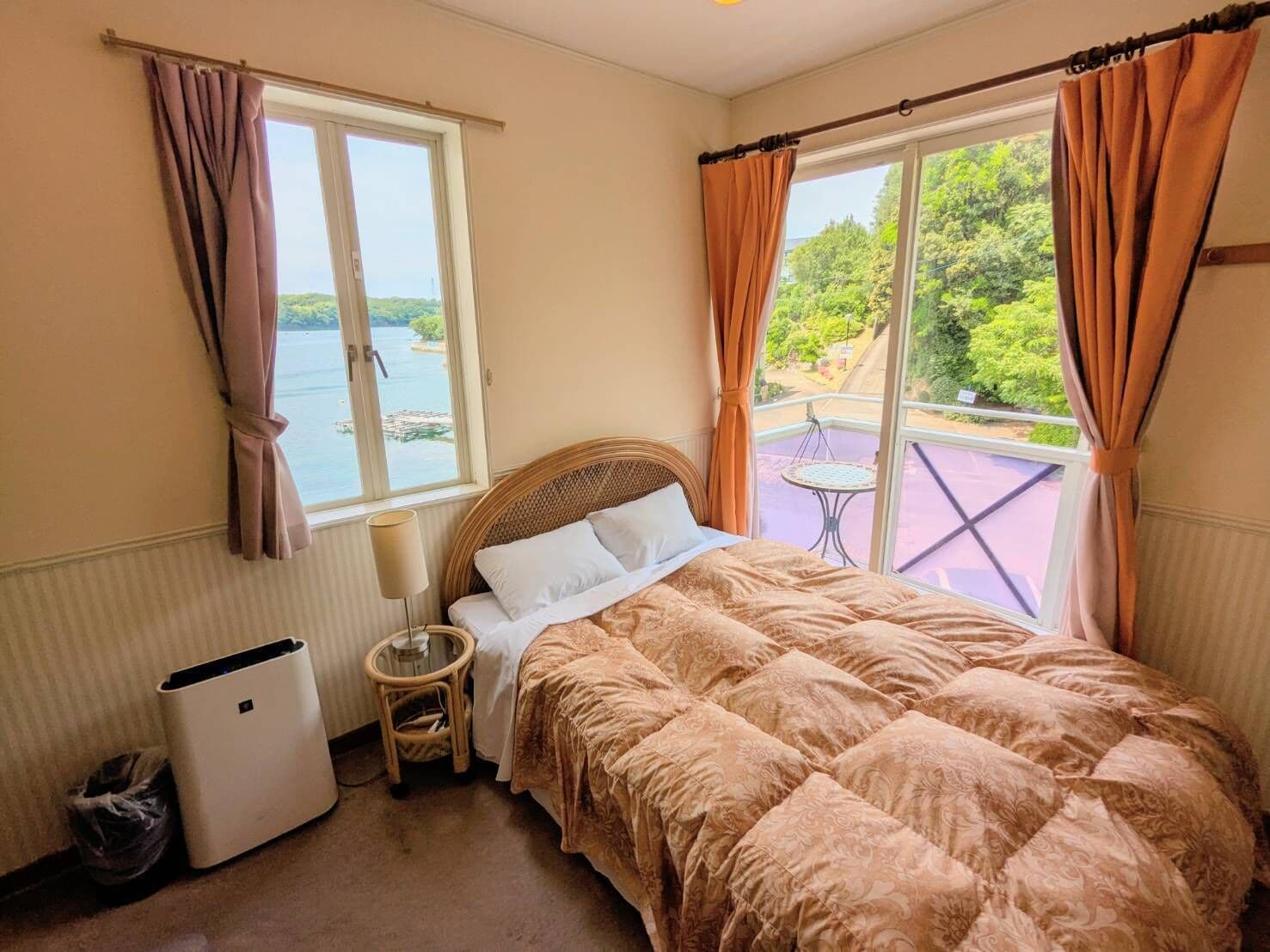 A bedroom with a bed, a small table, a waste bin, and a window with curtains.