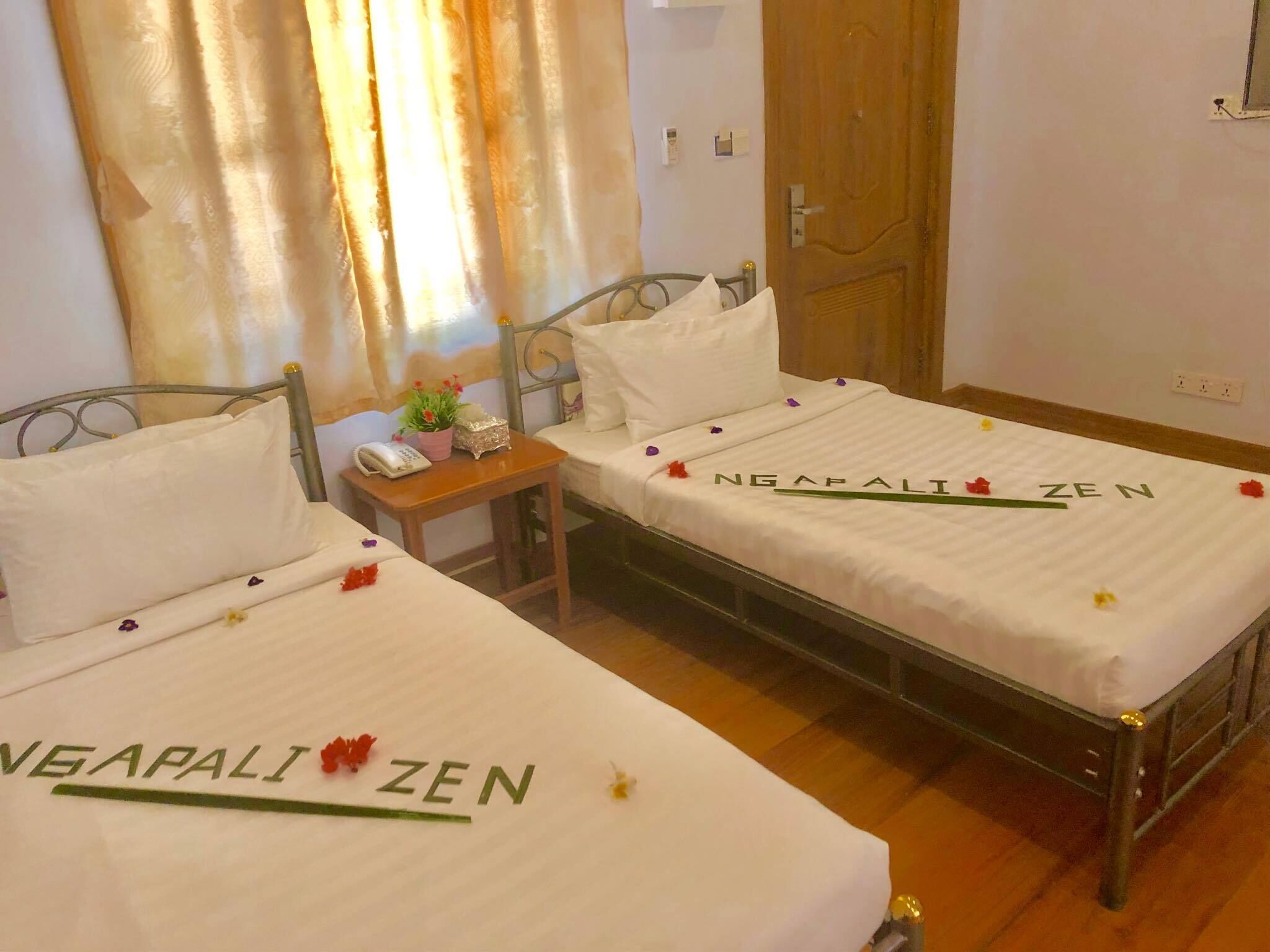 A hotel room with two beds, each bedspread has 'NEGAPALI ZEN' printed on it. There is a small table with a phone and a plant between the beds.