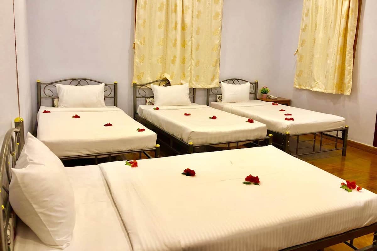 A hotel room with four single beds, each with white linens and red rose petals. The room has light-colored walls and curtains, and a small bedside table with a lamp.