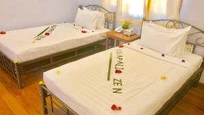 Dos camas individuales con ropa blanca y flores decorativas, una mesita de noche con un teléfono y una planta pequeña, y una ventana con cortinas.