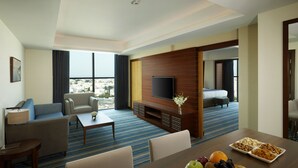 Una habitación de hotel moderna con un ventanal grande, un televisor de pantalla plana, una mesa de comedor con fruta y un sofá.
