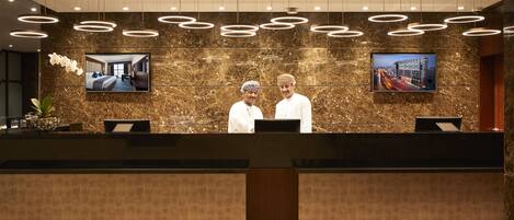 A reception desk with two staff members, a marble wall with two framed pictures, and a modern ceiling design.