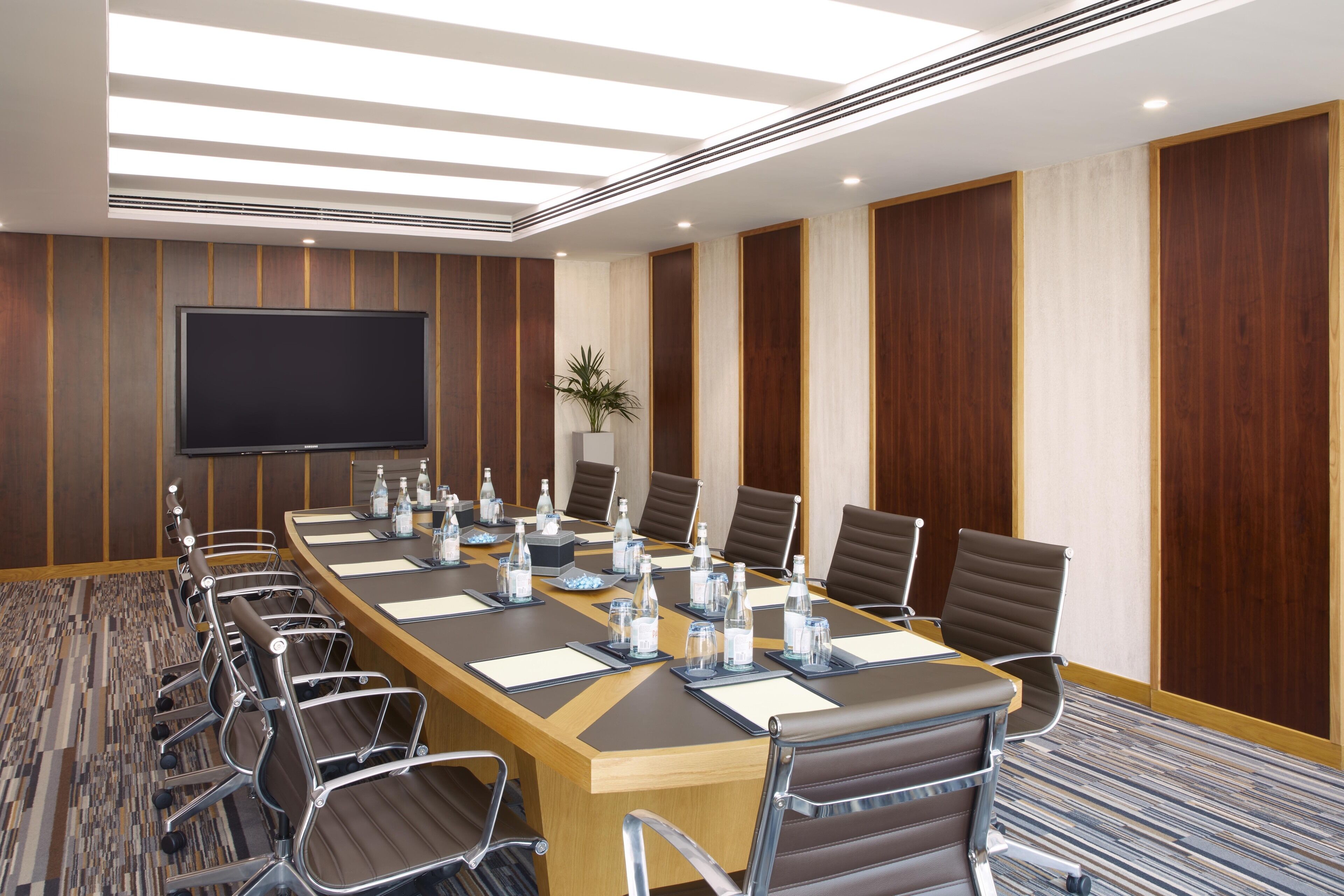 Une salle de conférence avec une longue table, de nombreuses chaises, un grand téléviseur à écran plat et une plante en pot.