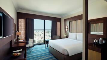 Una habitación de hotel con una cama grande, un escritorio con televisor, una silla, una mesita con fruta y vistas a la ciudad.