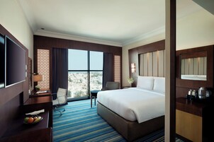 A hotel room with a large bed, a desk with a TV, a chair, a small table with fruit, and a view of the city.