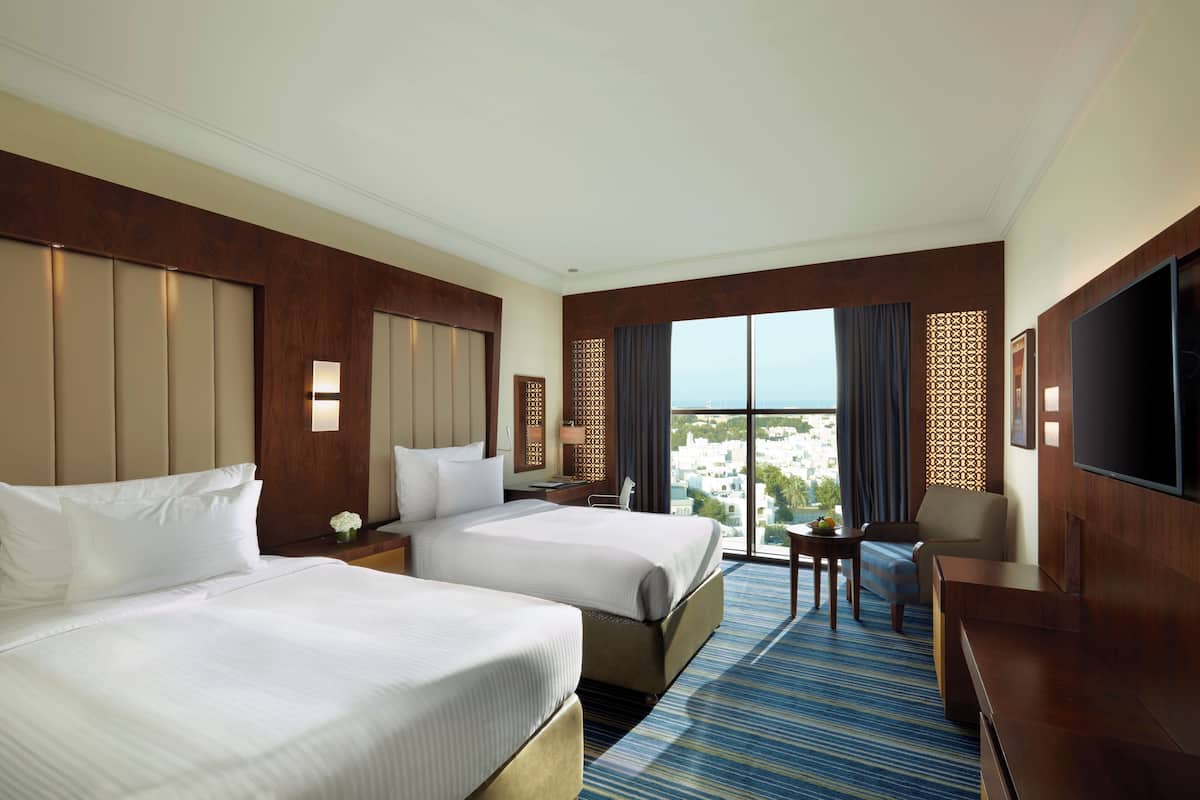 A hotel room with two beds, a desk, a chair, a TV, and a large window with a view.