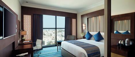 A hotel room with a large bed, a desk with a TV, a chair, a desk lamp, a coffee maker, and a view of the city.