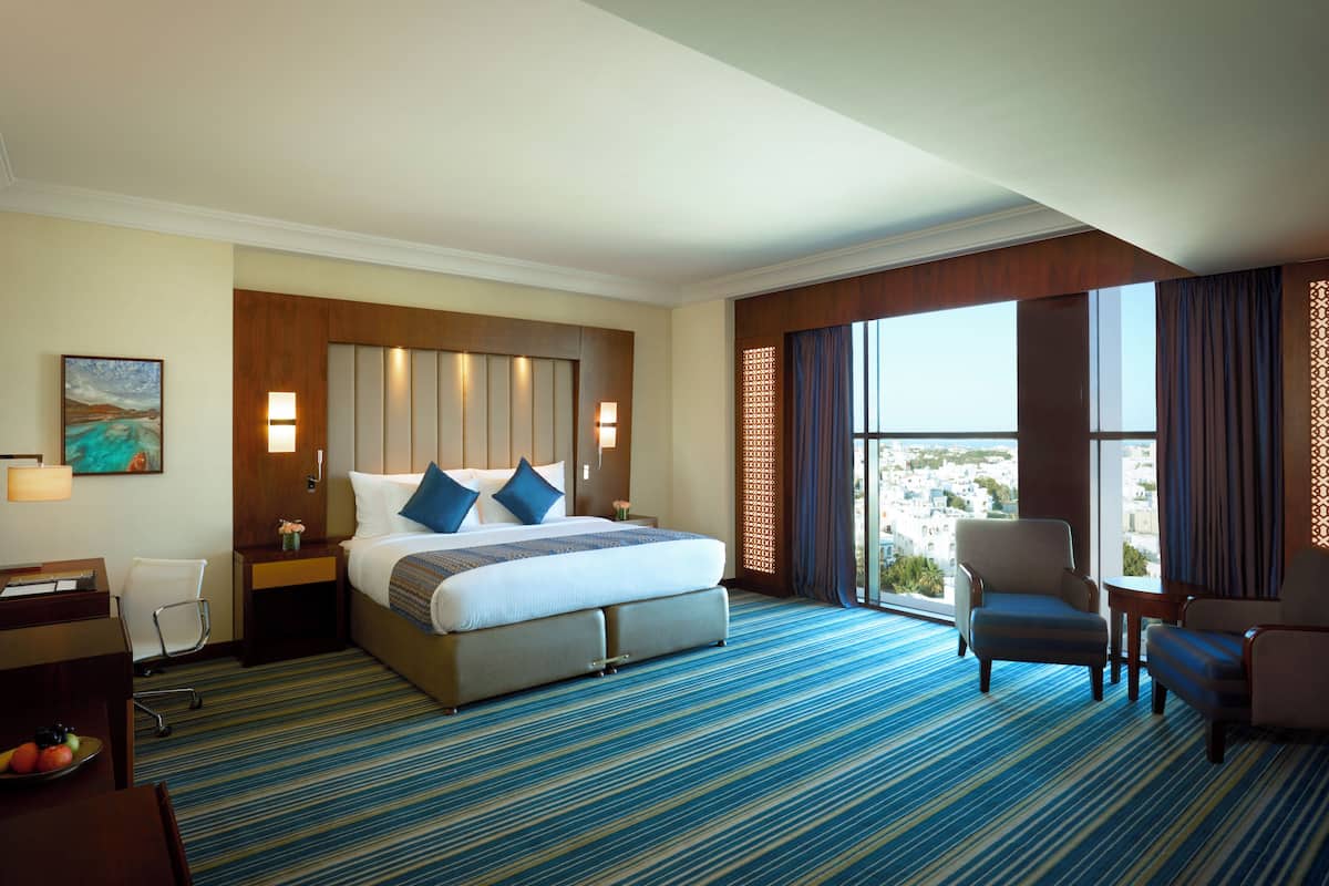 A hotel room with a large bed, two bedside tables, a desk, a chair, and a view of the city.