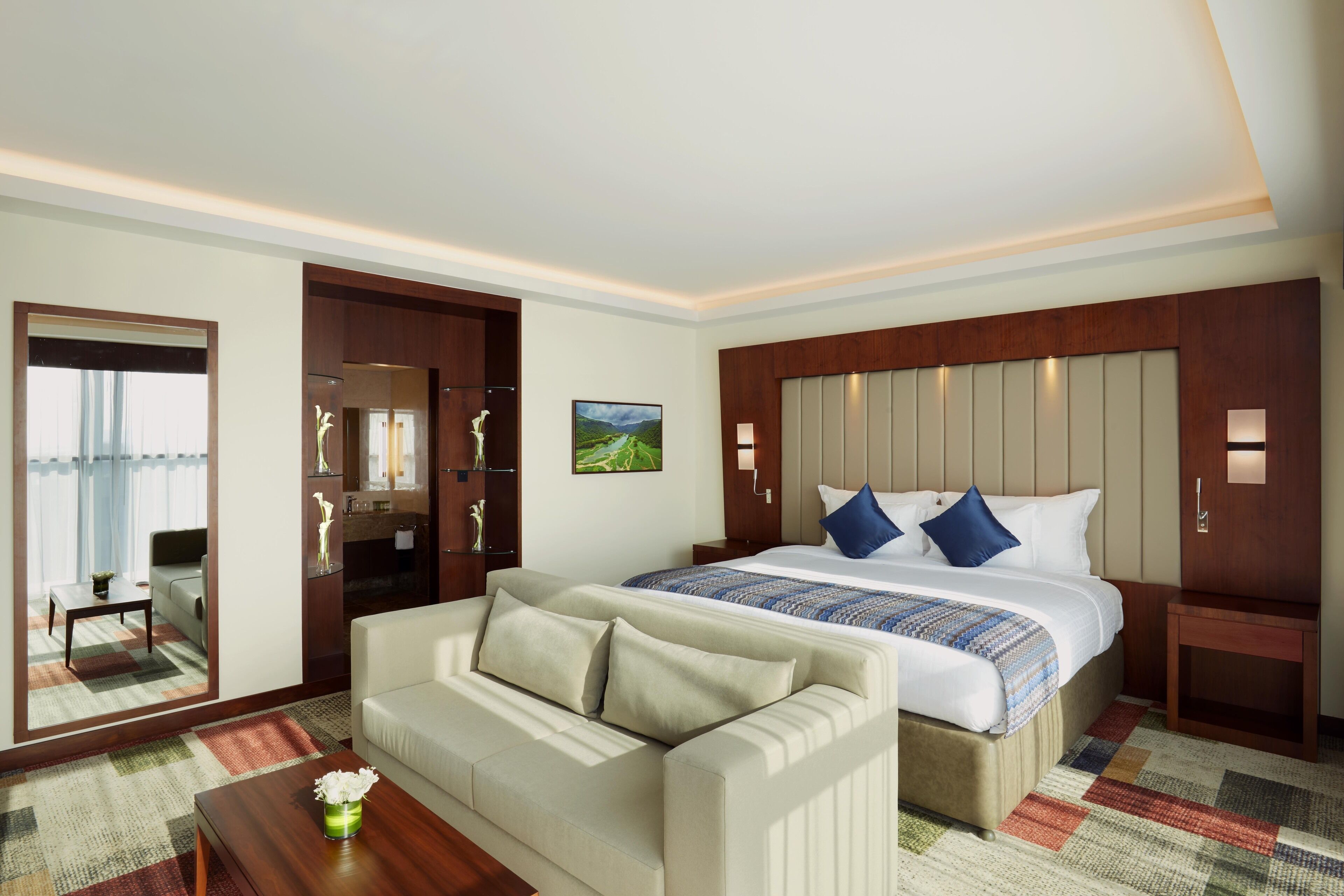 A hotel room with a large bed, a sofa, a small table with a vase, and a mirror.