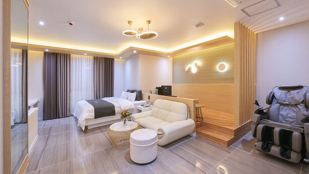 A modern hotel room with a bed, a sofa, a massage chair, a small table with a vase, and a wooden accent wall. - 1Beonji Muintel (Gwangyang)