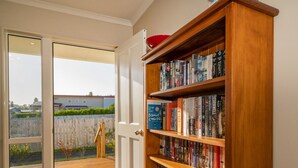 A room with a sliding glass door leading to a wooden deck, a bookshelf filled with books, and a red bowl on top. - Home Base - Cooks Beach Holiday Home (Cooks Beach)